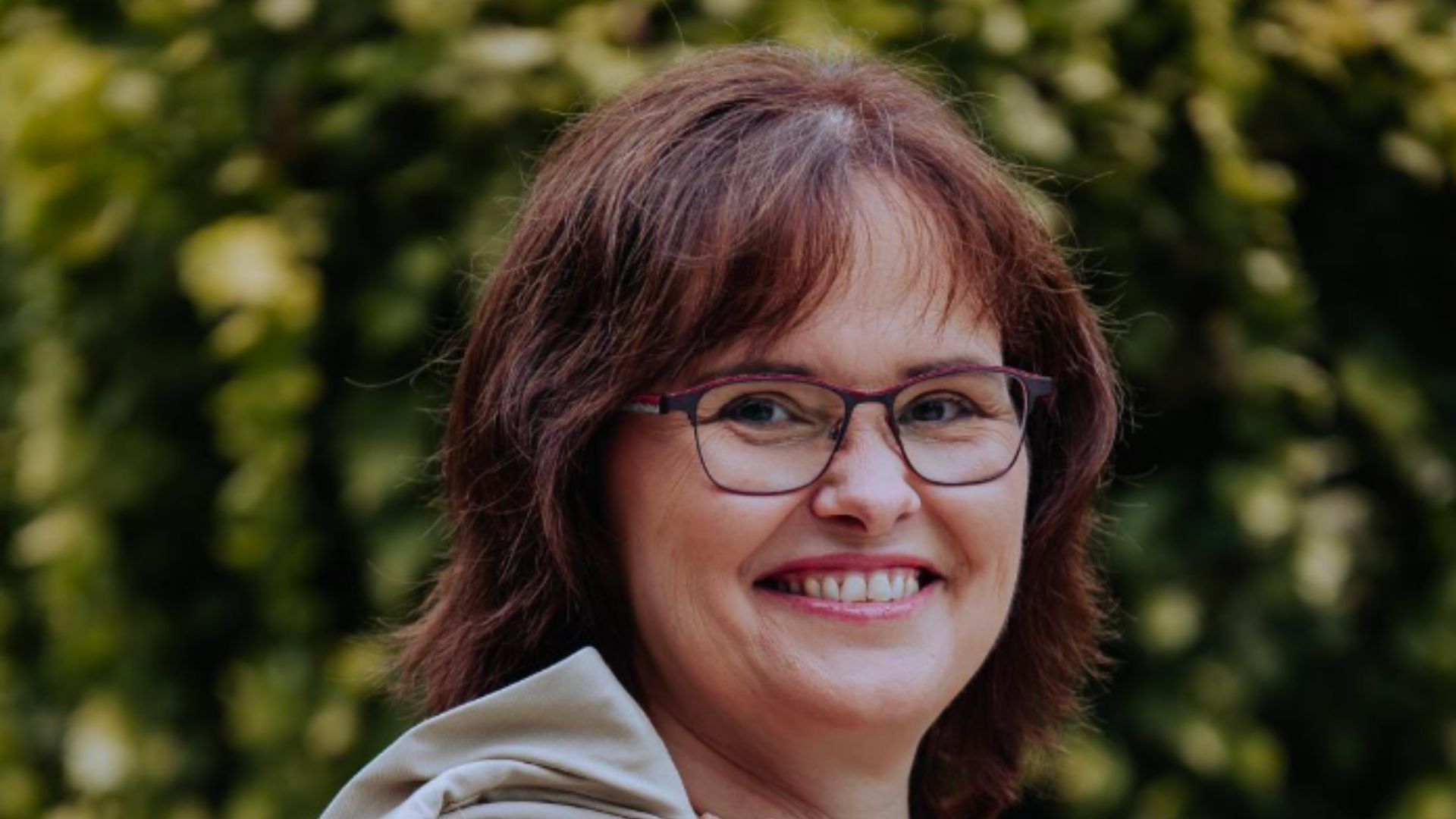 Frau mit Brille und braunen Haaren, die lächelt, vor einem grünen Hintergrund.