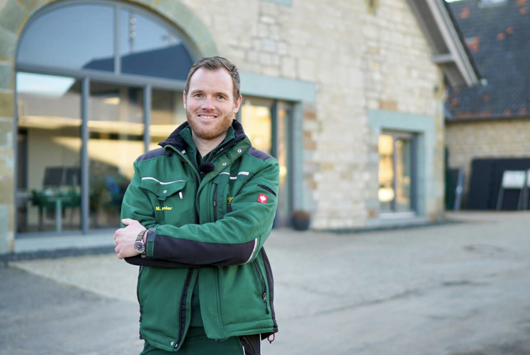 Mann in grüner Arbeitsjacke mit verschränkten Armen vor einem Gebäude mit großen Fenstern.